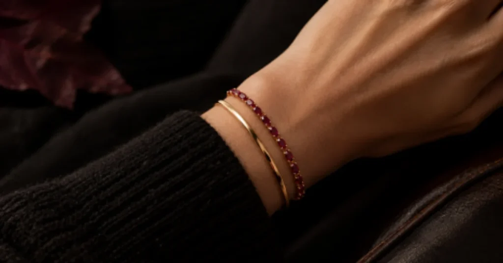 Close-up of a wrist wearing a red gemstone bracelet and a delicate gold bracelet beside a black knitted sleeve.